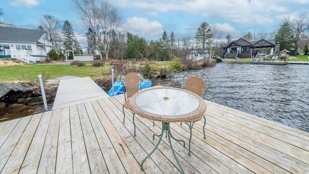 Um deck de madeira com uma mesa e cadeiras à beira de um rio, cercado por casas e árvores.