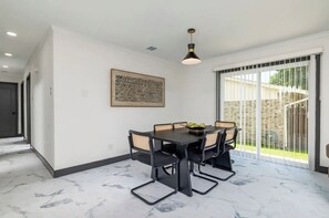 A dining area with a table, chairs, a pendant light, and a large window with a view of a brick wall and greenery.