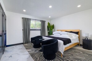 A modern hotel room with a bed, two chairs, a small table, a potted plant, and a window with curtains. - Home with Water Views Near
DFW, Arlington Stadiums (Irving)