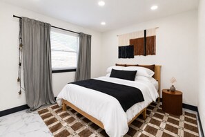 A bedroom with a bed, curtains, a wall-mounted artwork, a bedside table, and a patterned rug.