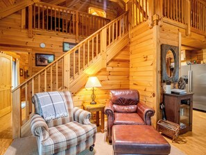 Ein gemütliches Blockhaus-Interieur mit einer Holztreppe, einem karierten Ohrensessel, einem Ledersessel, einem kleinen Tisch mit Lampe und einem Kühlschrank.