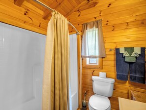 Et badeværelse i en træhytte med bruseforhæng, toilet og et vindue, hvor der hænger et håndklæde på væggen.