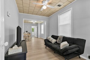 Ein Wohnzimmer mit schwarzem Sofa, Flachbildfernseher, Esstisch aus Holz und Deckenventilator.