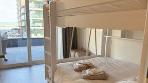 A bunk bed with a white mattress and pillows, a small table with a chair, and a view of a building through a glass door.