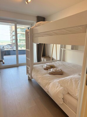 Ein Etagenbett mit weißer Matratze und Kissen, ein kleiner Tisch mit Stuhl sowie Blick auf ein Gebäude durch eine Glastür.