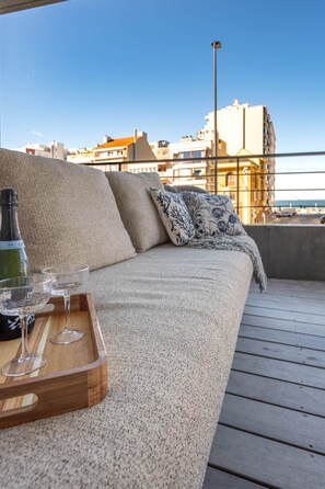 En balkon med en sofa, et bakke med glas og en flaske, og et bybillede i baggrunden.