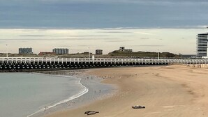 A beach with a rocky foreground, a sandy shore, and a pier extending into the water.