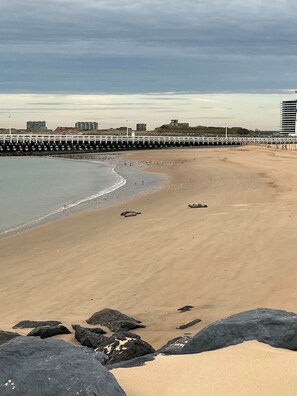 Ein Strand mit felsigem Vordergrund, einem sandigen Ufer und einem Pier, der sich ins Wasser erstreckt.