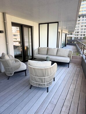 Ein Balkon mit einer Couch und Korbstühlen, eine Schiebetür und Blick auf Gebäude.