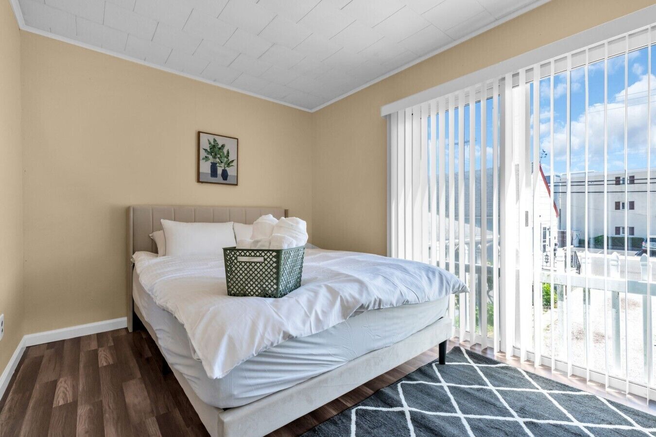 A bedroom with a bed, a basket of towels, a framed picture, and a view through large windows.