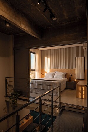 A modern bedroom with a large bed, a bedside table, a nightstand, a window with curtains, and a staircase with a railing.