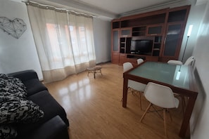 A living room with a sofa, a glass-top dining table, white chairs, a TV, and a wooden cabinet. - 1A154 Gijón like at home Apartment for 5 pax (Gijón)