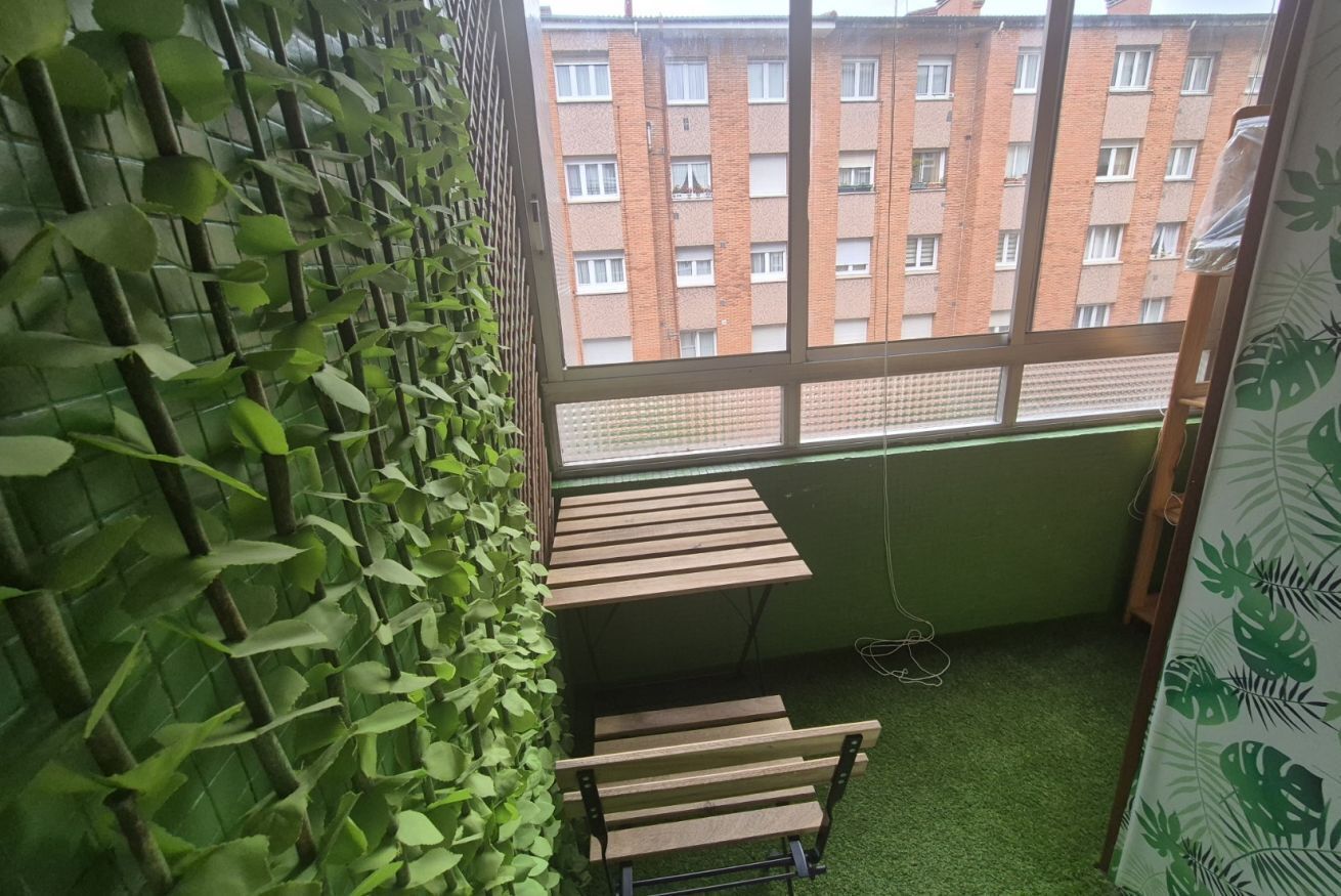 A room with a green leafy wall, a window with a view of a brick building, and a small wooden table.