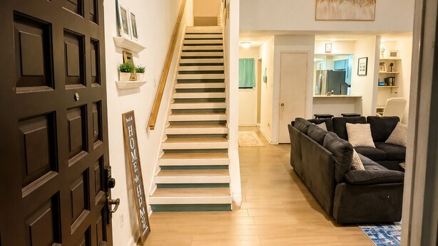 A staircase leading to a second floor, a living room with a dark-colored sofa, a kitchen area, and a patterned rug on the floor. - Modern 3 Story Home 9 Min to DFW w King Suite Pool (Euless)