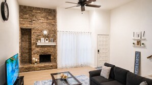 A living room with a sofa, coffee table, TV, fireplace, and a ceiling fan. - Modern 3 Story Home 9 Min to DFW w King Suite Pool (Euless)