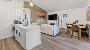 Ett modernt kök med en köksö i mitten, en matplats med ett träbord och stolar, en soffa och en platt-TV som är monterad på väggen.