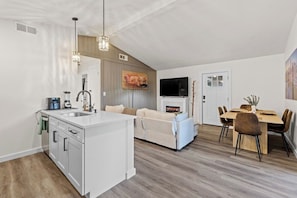 A modern kitchen with a central island, a dining area with a wooden table and chairs, a sofa, and a flat-screen TV mounted on the wall. - Cozy Austin Getaway | Game Room + Large Backyard (Austin)