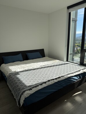 Un dormitorio moderno con una cama grande, vistas de un paisaje urbano a través de una ventana y un cabecero oscuro.