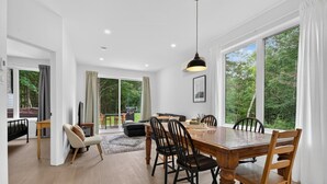 A spacious dining area with a wooden table and chairs, large windows with a view of greenery, and a ceiling light fixture. - Le Relais Austin 4 — Spa, Nature & Adventure (Austin)