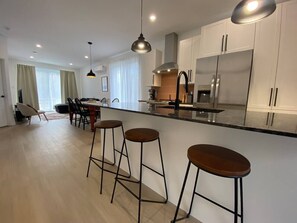 Una cocina moderna con una isla central, taburetes de bar, iluminación colgante y electrodomésticos de acero inoxidable.