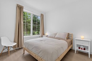 Un dormitorio con una cama de madera, mesitas de noche blancas, una silla blanca y una ventana con cortinas.