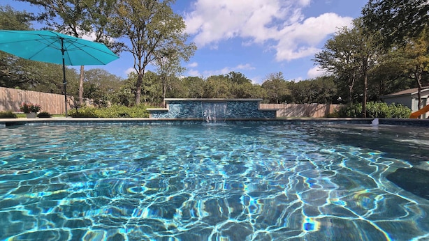 A clear swimming pool with a waterfall feature, surrounded by trees and a wooden fence. - Gruene Gables Pool & Game Room (New Braunfels)