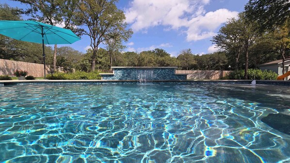 A clear swimming pool with a waterfall feature, surrounded by trees and a wooden fence. - Gruene Gables Pool & Game Room (New Braunfels)