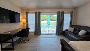 A room with a desk, a black leather sofa, a TV mounted on the wall, and a sliding glass door leading to an outdoor area with a pool and chairs. - Gruene Gables Pool & Game Room (New Braunfels)