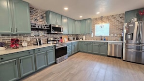 A kitchen with teal cabinets, a brick wall, a stainless steel refrigerator, and a microwave. - Gruene Gables Pool & Game Room (New Braunfels)