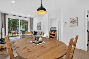 A modern dining area with a round wooden table, chairs, a pendant light, and a view of the outdoors.
