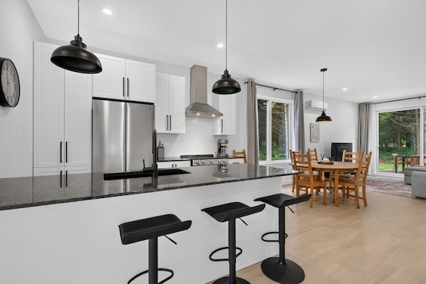 A modern kitchen with white cabinets, a stainless steel refrigerator, a dining area with wooden chairs, and a bar with stools.