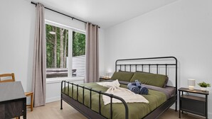 A modern bedroom with a metal bed frame, a bedside table with a lamp, and a view of trees through a window.