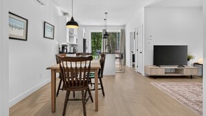 Ett modernt kök med vita skåp, en matplats med ett träbord och stolar, en tv på ett träställ och ett stort fönster med utsikt över grönområden.