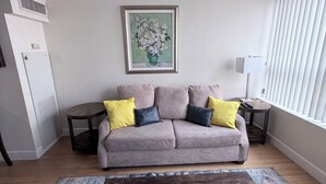A living room with a grey sofa, yellow and dark grey cushions, a framed floral artwork, a small round table with a lamp, and a window with blinds. - Cozy 1-bedroom condo in serene Reno with fitness room and AC (Reno)