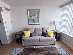 Ein Wohnzimmer mit grauem Sofa, gelben und dunkelgrauen Kissen, einem gerahmten Blumenbild, einem kleinen runden Tisch mit Lampe und einem Fenster mit Jalousien.