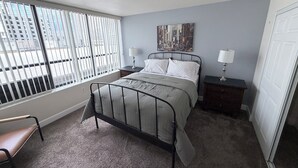 A bedroom with a bed, two bedside tables with lamps, a window with blinds, a chair, and a door. - Cozy 1-bedroom condo in serene Reno with fitness room and AC (Reno)