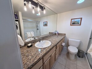 Ein Badezimmer mit einer Granitplatte, einem weißen Waschbecken, einer Toilette, einem Mülleimer und einer Dusche.