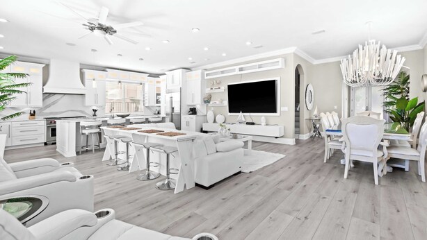 A modern living room with a large TV, white furniture, and a kitchen area with white cabinets. - Miramar Beach Home, Pool, Hot Tub & Chef's Kitchen (Miramar Beach)