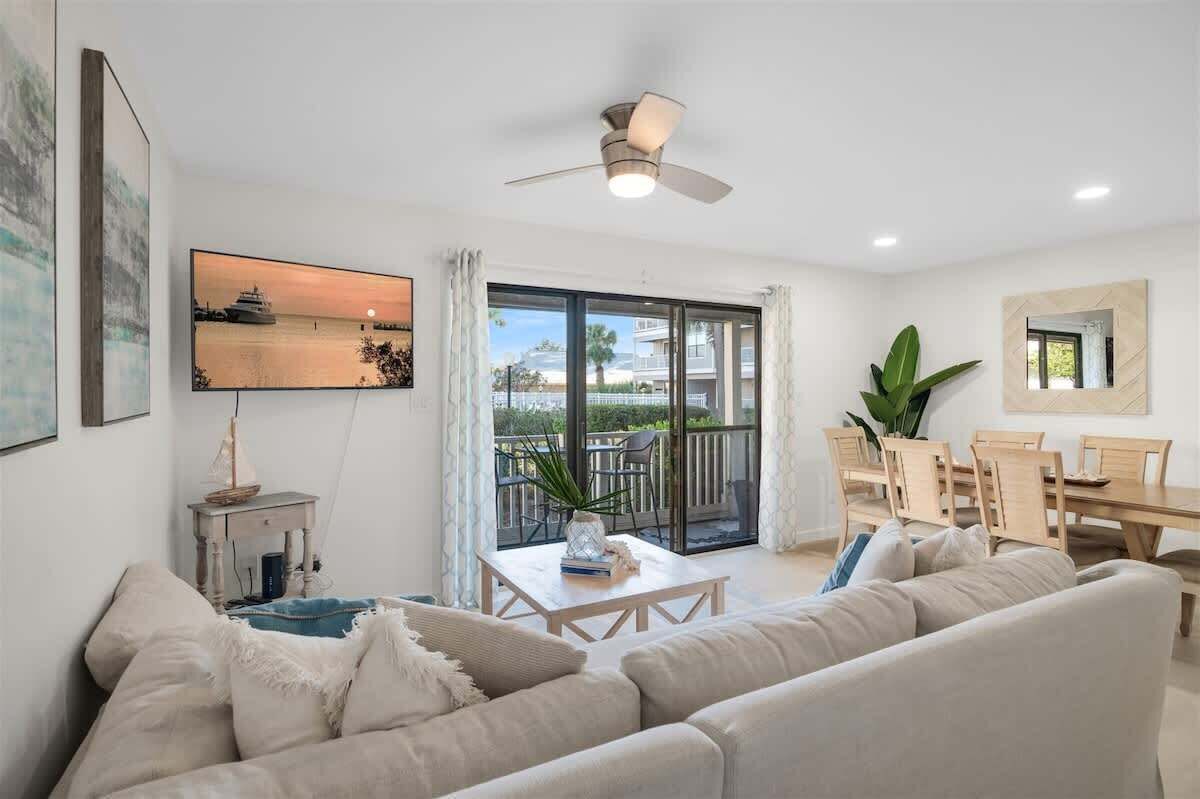 A modern living room with a sofa, a coffee table, and a dining area with chairs and a table. There is a balcony with a view of the sea and a ceiling fan.