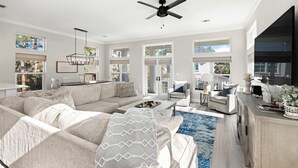 A spacious living room with a large sofa, a coffee table, and a ceiling fan. There are large windows providing natural light and a view of the outdoors. - Santa Rosa Home with Private Pool, Near Beach (Santa Rosa Beach)