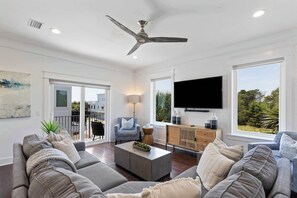 A modern living room with a ceiling fan, a flat-screen TV, and a large window offering a view of greenery. - Inlet Beach Home with Shared Pool, Cart & Bikes (Inlet Beach)