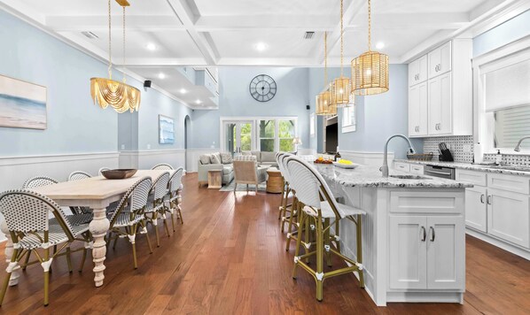 A modern kitchen with white cabinetry, a marble countertop, and a dining area with wicker chairs and a wooden table. - Seacrest Home with Pool & Private Beach Access (Rosemary Beach)