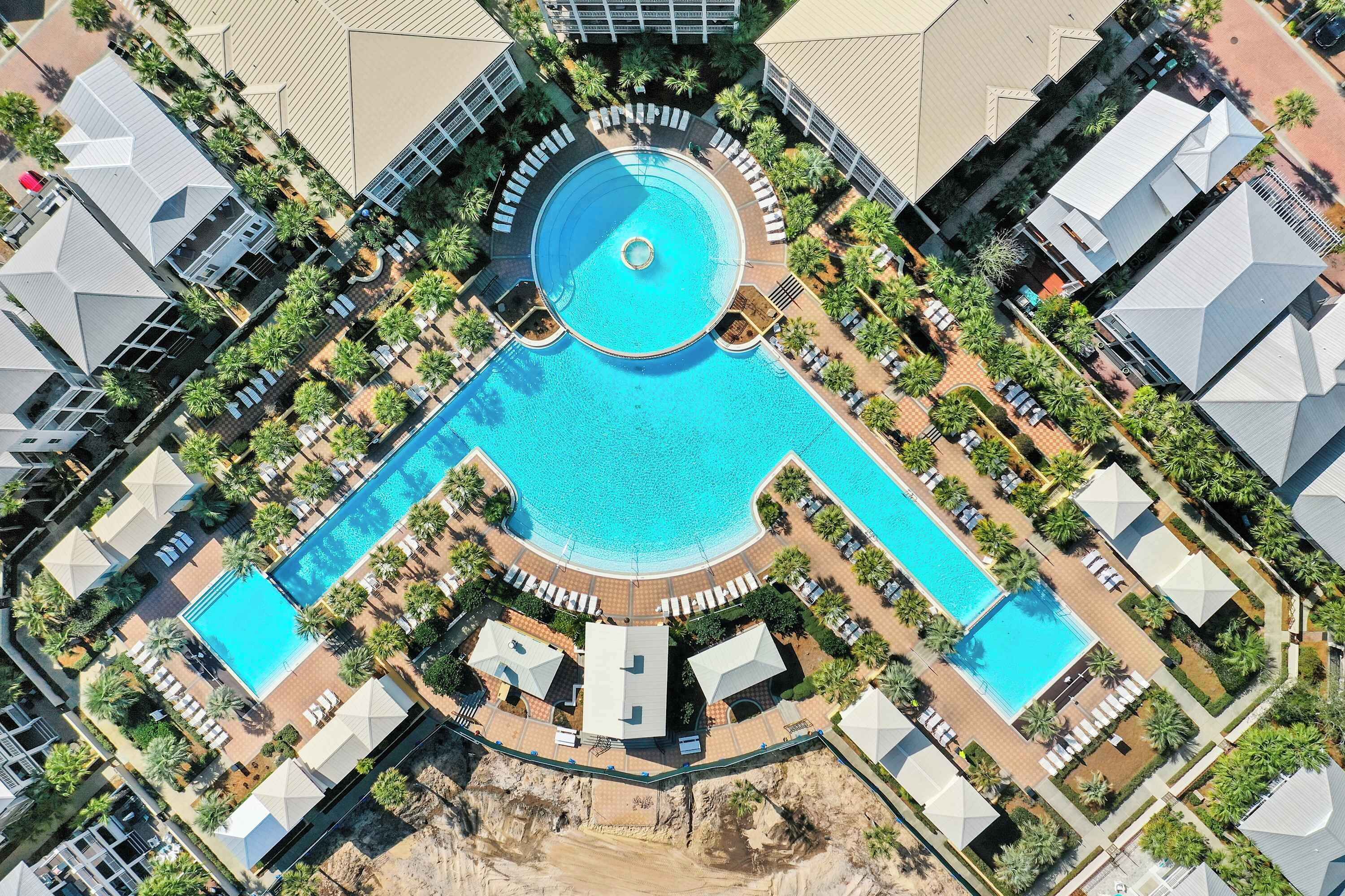 Vista aérea de un complejo turístico con una piscina central, varias zonas de descanso y palmeras a su alrededor.