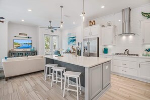 Eine moderne Küche mit weißen Schränken, einer großen Kochinsel mit Hockern, einem Deckenventilator und einem an der Wand montierten Fernseher.