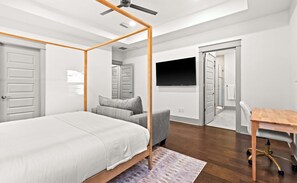 Ein Schlafzimmer mit Himmelbett, grauem Sofa, Schreibtisch aus Holz und einem an der Wand montierten Fernseher.