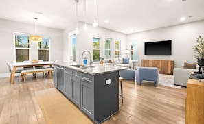 Eine moderne Küche mit Kochinsel, ein Essbereich mit Tisch und Stühlen, ein Wohnzimmer mit Sofa und Sesseln sowie ein an der Wand befestigter Flachbildfernseher.