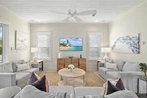 Ein Wohnzimmer mit Deckenventilator, zwei Sofas, einem Couchtisch und einem Fernseher, der eine Strandansicht zeigt.