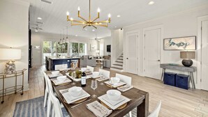 Une salle à manger spacieuse avec une grande table en bois, dressée pour six personnes, des chaises blanches et un lustre.