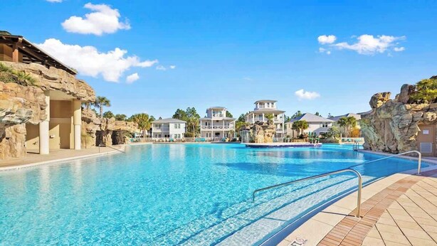 Uma piscina com águas azul-claras, rodeada por edifícios com varandas e uma paisagem rochosa.
