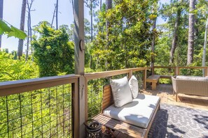 Eine Holzterrasse mit einer Couch und einem Korbstuhl, umgeben von einem Metallzaun und üppiger Bepflanzung.